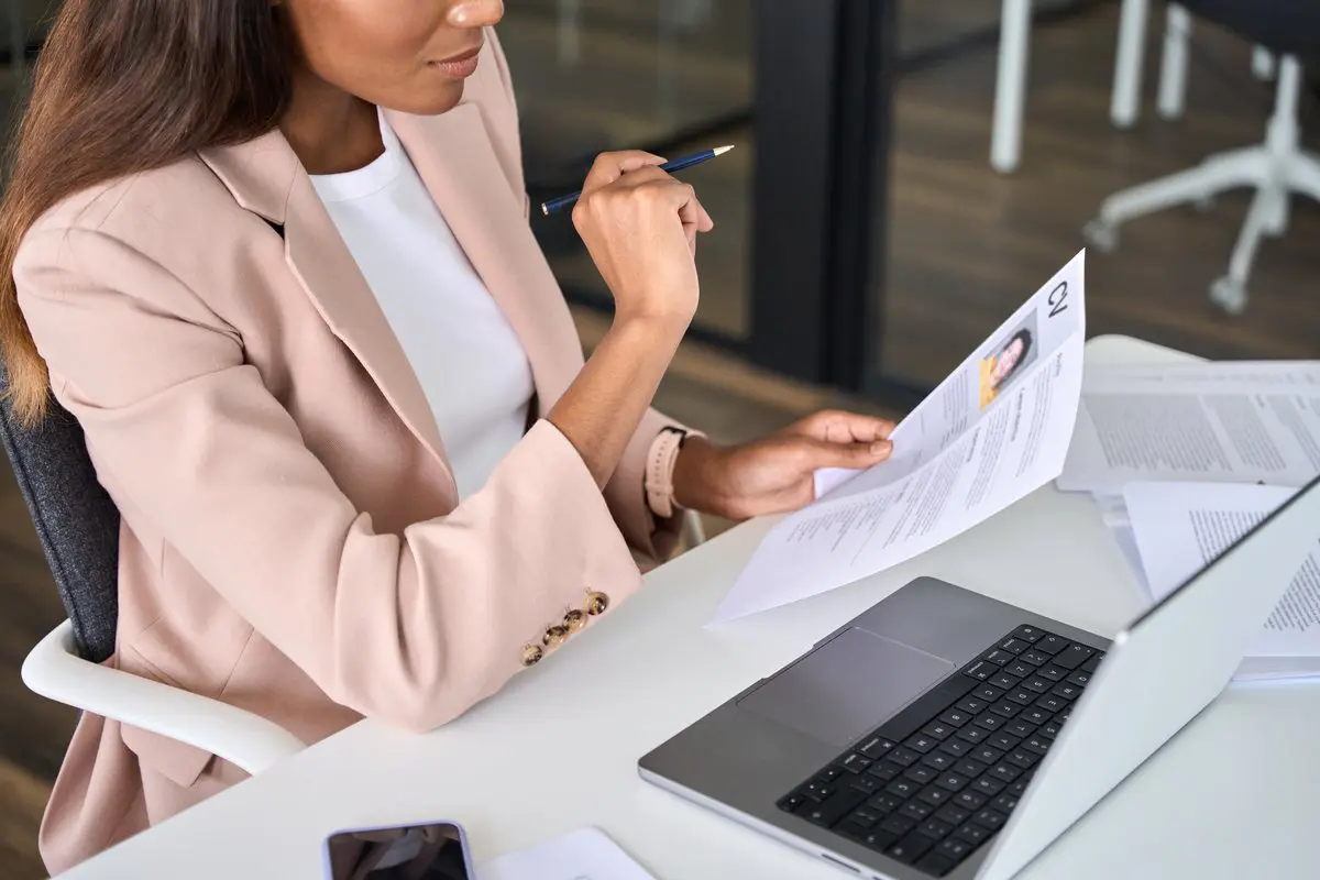 Updating your resume A woman in a blazer reviewing and updating her resume at a desk with a laptop, preparing for a new job search