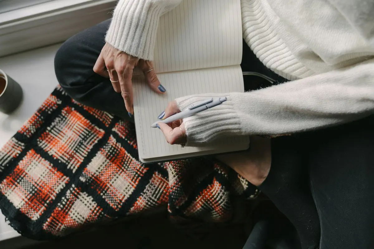 Gratitude journaling A woman wrapped in a cozy sweater and plaid blanket writing in a journal with a warm drink by a window, practicing gratitude journaling to calm the mind